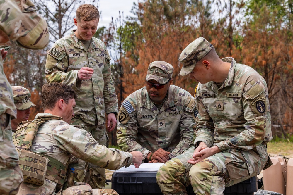 A group of U.S. Army soldiers in OCP (Operational Camouflage Pattern) uniforms are gathered outdoors in a wooded area, reviewing documents or maps on a hard case surface. Visible name tapes identify at least three soldiers: SMITH, JACKSON, and KIMBALL. The scene appears to be a field planning or briefing session during a training exercise or field operation.