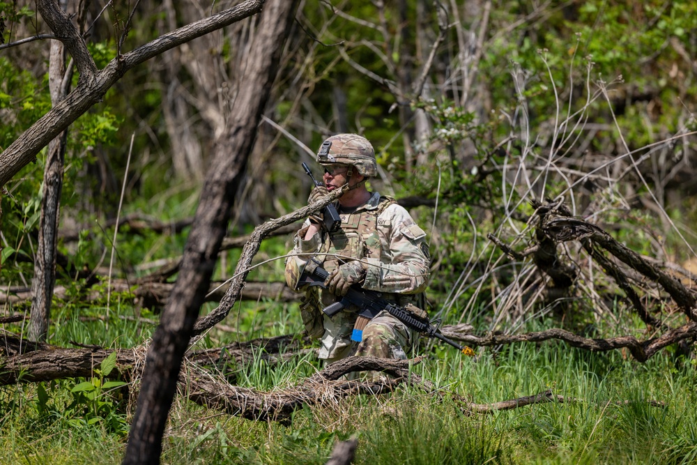 A soldier in full camouflage gear is positioned in a wooded area among fallen branches and undergrowth, appearing to communicate via radio while holding a rifle. The soldier is wearing MultiCam/OCP pattern uniform with a tactical vest, helmet, and gloves. A blue training marker is visible on the rifle, suggesting this is a military training exercise rather than a combat situation.