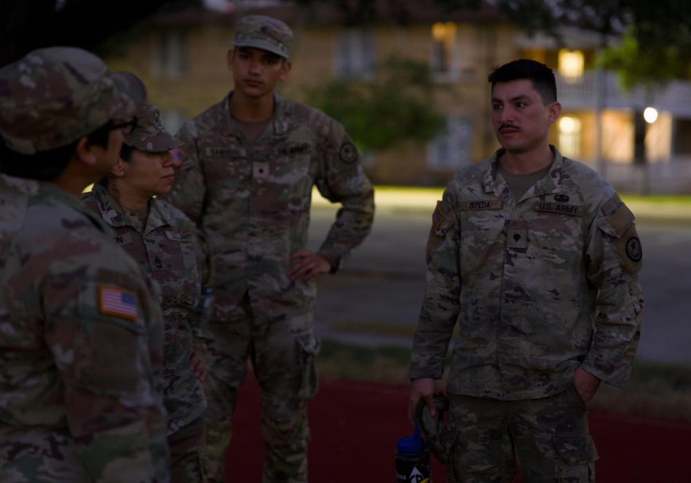 Four U.S. Army soldiers in OCP (Operational Camouflage Pattern) uniforms are gathered in an outdoor area during dusk or early morning, engaged in conversation. The soldier on the right is identified by a name tape reading 'ZEPEDA' and a 'U.S. ARMY' tape, while others display American flag patches on their shoulders. The setting appears to be a military installation with buildings and trees visible in the background.
