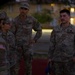 Four U.S. Army soldiers in OCP (Operational Camouflage Pattern) uniforms are gathered in an outdoor area during dusk or early morning, engaged in conversation. The soldier on the right is identified by a name tape reading 'ZEPEDA' and a 'U.S. ARMY' tape, while others display American flag patches on their shoulders. The setting appears to be a military installation with buildings and trees visible in the background.