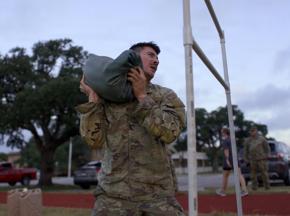 A soldier in MultiCam/OCP camouflage uniform is carrying a heavy sandbag or ruck sack on his shoulder, appearing to be engaged in a physical fitness or training exercise. The setting appears to be a military installation or base, likely during early morning or evening hours given the overcast, dim lighting. Additional personnel in military and civilian attire are visible in the background.