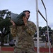A soldier in MultiCam/OCP camouflage uniform is carrying a heavy sandbag or ruck sack on his shoulder, appearing to be engaged in a physical fitness or training exercise. The setting appears to be a military installation or base, likely during early morning or evening hours given the overcast, dim lighting. Additional personnel in military and civilian attire are visible in the background.