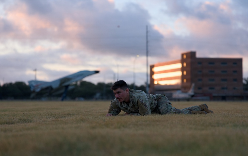 A military service member in camouflage uniform is performing a low crawl exercise on a grass field during dusk or dawn. The setting appears to be a military installation, evidenced by a static display aircraft and a multi-story brick building visible in the background. The warm sunset light illuminates the building's windows, creating a dramatic backdrop.