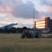 A military service member in camouflage uniform is performing a low crawl exercise on a grass field during dusk or dawn. The setting appears to be a military installation, evidenced by a static display aircraft and a multi-story brick building visible in the background. The warm sunset light illuminates the building's windows, creating a dramatic backdrop.