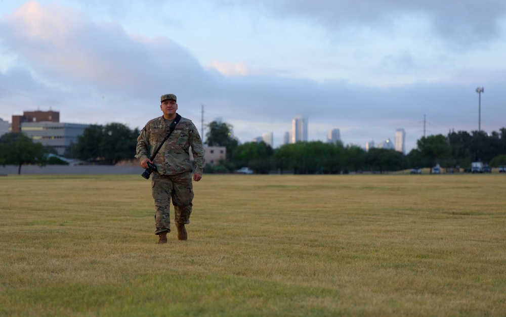 A military service member in MultiCam/OCP camouflage uniform walks across a large open grass field carrying a camera with a shoulder strap. A city skyline is visible in the background, suggesting proximity to an urban area. The photo is taken during early morning or evening hours under a partly cloudy sky.
