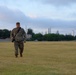 A military service member in MultiCam/OCP camouflage uniform walks across a large open grass field carrying a camera with a shoulder strap. A city skyline is visible in the background, suggesting proximity to an urban area. The photo is taken during early morning or evening hours under a partly cloudy sky.