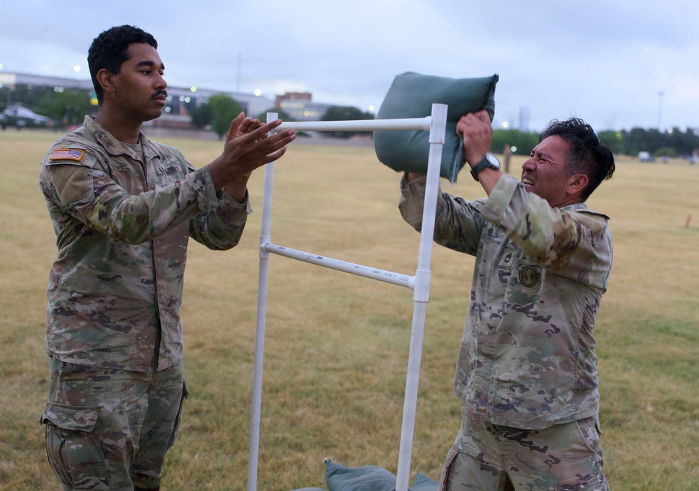 Two U.S. Army soldiers in OCP (Operational Camouflage Pattern) uniforms are engaged in a physical training or competition activity on a grassy field. One soldier is lifting or placing a green sandbag onto a white PVC pipe rack structure while the other observes with hands raised, possibly spotting or judging. The activity appears to be part of a military fitness event or challenge.