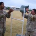 Two U.S. Army soldiers in OCP (Operational Camouflage Pattern) uniforms are engaged in a physical training or competition activity on a grassy field. One soldier is lifting or placing a green sandbag onto a white PVC pipe rack structure while the other observes with hands raised, possibly spotting or judging. The activity appears to be part of a military fitness event or challenge.