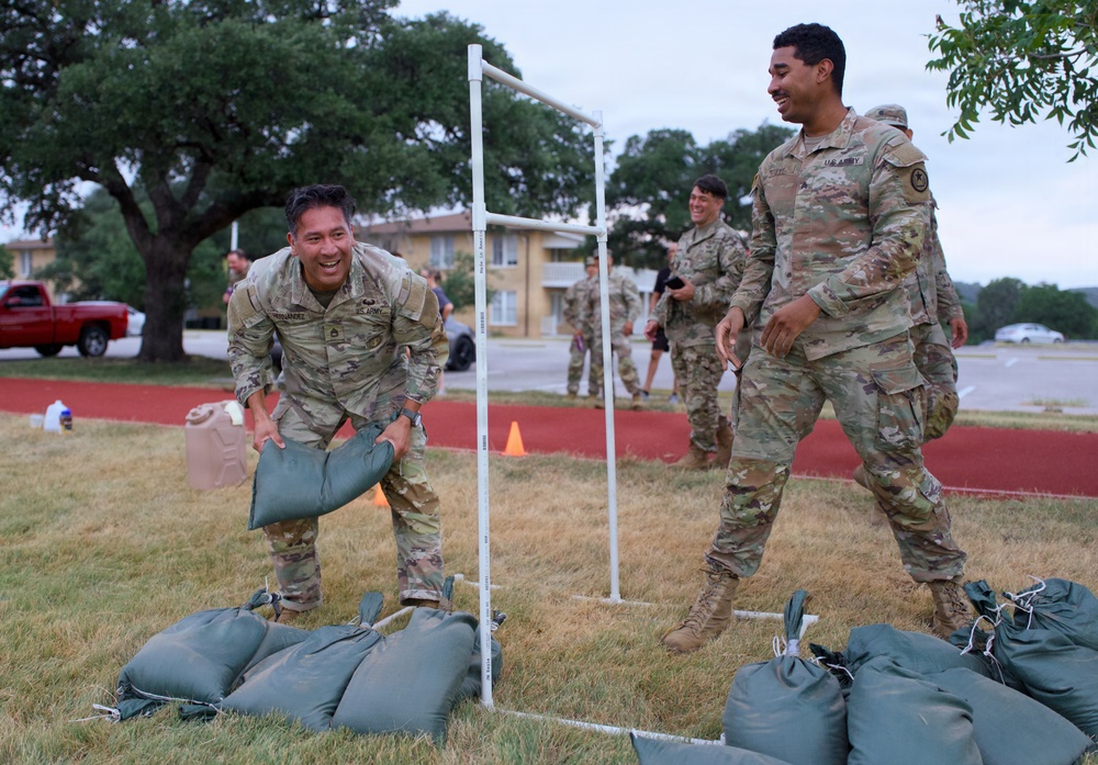 U.S. Army soldiers in OCP (Operational Camouflage Pattern) uniforms are engaged in what appears to be a physical fitness or team competition event on a military installation. Two soldiers in the foreground are smiling and handling green sandbags near a white PVC pipe goal structure, suggesting a competitive exercise or training activity. Additional soldiers are visible in the background observing or participating.