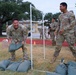 U.S. Army soldiers in OCP (Operational Camouflage Pattern) uniforms are engaged in what appears to be a physical fitness or team competition event on a military installation. Two soldiers in the foreground are smiling and handling green sandbags near a white PVC pipe goal structure, suggesting a competitive exercise or training activity. Additional soldiers are visible in the background observing or participating.
