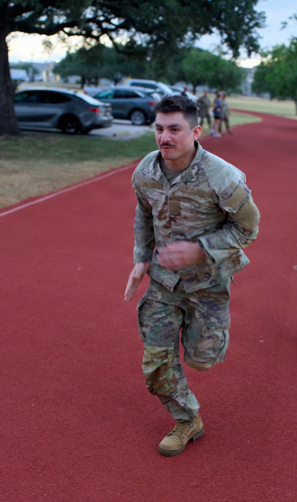 A male soldier in US Army OCP (Operational Camouflage Pattern) uniform is running on a red rubberized track, carrying what appears to be a rucksack or gear on his back. The setting appears to be a military installation fitness track during early morning or evening hours. Other personnel in uniform are visible in the background near a parking area.