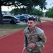 A male soldier in US Army OCP (Operational Camouflage Pattern) uniform is running on a red rubberized track, carrying what appears to be a rucksack or gear on his back. The setting appears to be a military installation fitness track during early morning or evening hours. Other personnel in uniform are visible in the background near a parking area.