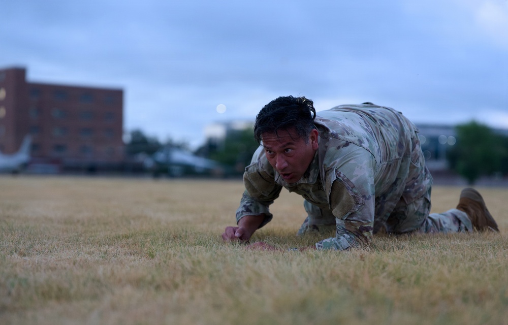 A military service member in Operational Camouflage Pattern (OCP) uniform is crawling low across a dry grass field, likely performing a low-crawl exercise or physical training drill. The image is taken at dusk or dawn, with blurred buildings and trees visible in the background. The subject appears focused and physically exerted.