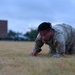 A military service member in Operational Camouflage Pattern (OCP) uniform is crawling low across a dry grass field, likely performing a low-crawl exercise or physical training drill. The image is taken at dusk or dawn, with blurred buildings and trees visible in the background. The subject appears focused and physically exerted.