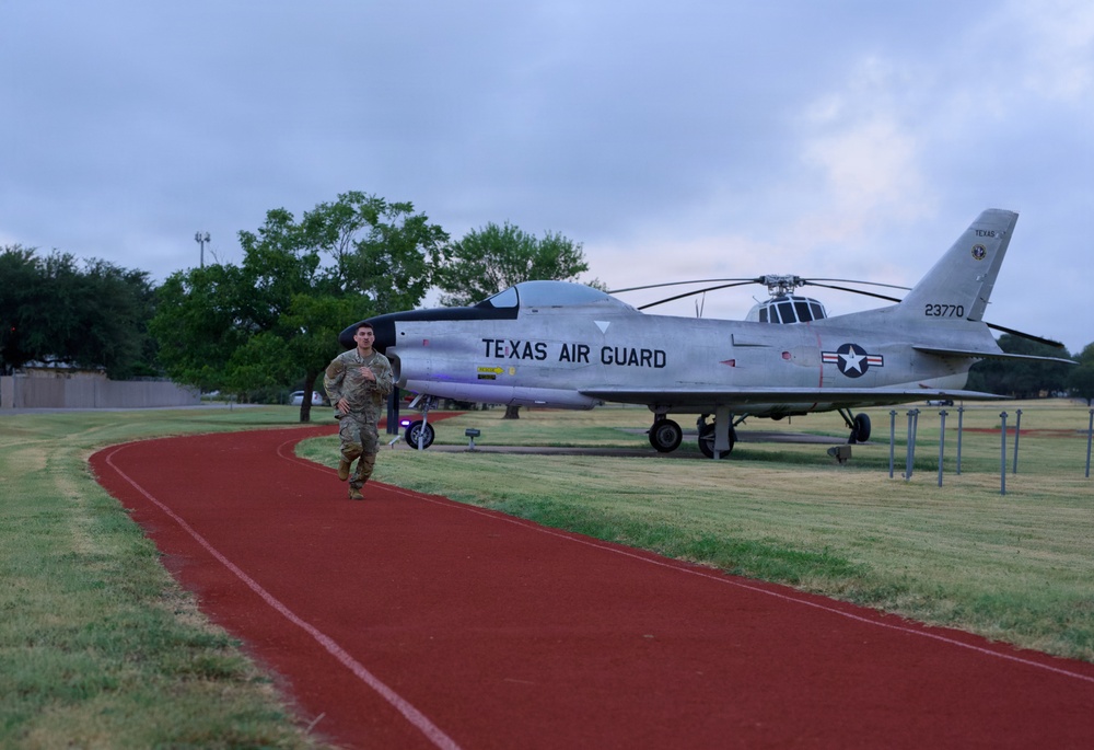 A military service member in camouflage uniform is running on a red track at what appears to be a Texas Air National Guard base. A static display aircraft, marked 'TEXAS AIR GUARD' with serial number 23770, is prominently positioned on the grass beside the track. The setting appears to be early morning or evening based on the overcast, dim lighting conditions.