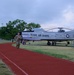 A military service member in camouflage uniform is running on a red track at what appears to be a Texas Air National Guard base. A static display aircraft, marked 'TEXAS AIR GUARD' with serial number 23770, is prominently positioned on the grass beside the track. The setting appears to be early morning or evening based on the overcast, dim lighting conditions.