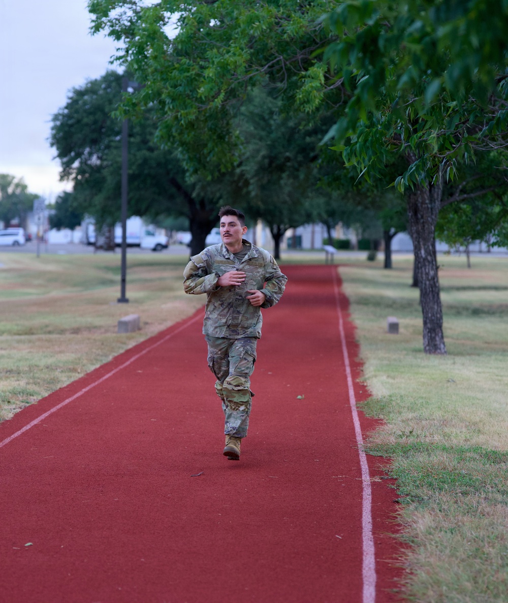 A military service member in full camouflage uniform and combat boots is running on a red rubberized track, likely during a physical fitness test or training exercise. The setting appears to be a military installation, with manicured grounds, lamp posts, and parked vehicles visible in the background. The photo is taken during early morning or overcast conditions.