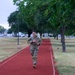 A military service member in full camouflage uniform and combat boots is running on a red rubberized track, likely during a physical fitness test or training exercise. The setting appears to be a military installation, with manicured grounds, lamp posts, and parked vehicles visible in the background. The photo is taken during early morning or overcast conditions.