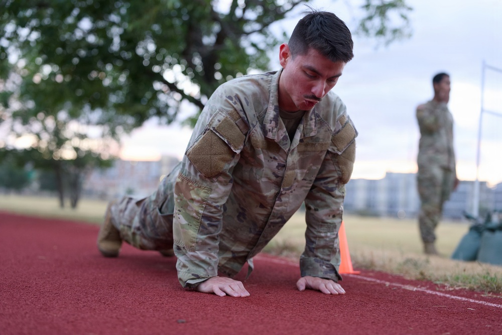 A soldier in Operational Camouflage Pattern (OCP) uniform is performing push-ups on a red rubberized running track. A second soldier in matching uniform is visible in the blurred background, standing and observing. The setting appears to be a military installation fitness area during early morning or evening hours.