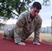 A soldier in Operational Camouflage Pattern (OCP) uniform is performing push-ups on a red rubberized running track. A second soldier in matching uniform is visible in the blurred background, standing and observing. The setting appears to be a military installation fitness area during early morning or evening hours.