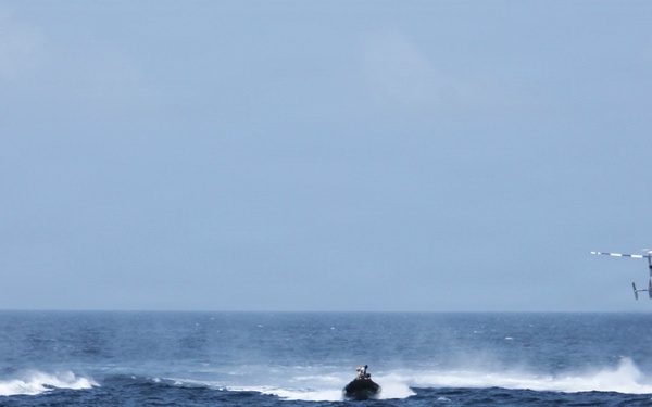 The image shows a maritime operation involving three fast-moving watercraft and one helicopter over open ocean. The vessels are traveling at high speed, generating significant wake and spray. This appears to be a coordinated naval or coast guard exercise or interdiction operation.