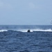The image shows a maritime operation involving three fast-moving watercraft and one helicopter over open ocean. The vessels are traveling at high speed, generating significant wake and spray. This appears to be a coordinated naval or coast guard exercise or interdiction operation.
