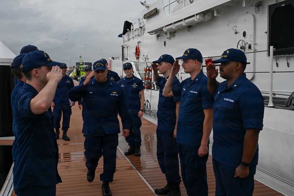 A group of U.S. Coast Guard personnel are gathered on the deck of a white Coast Guard cutter, performing a formal salute ceremony. A senior officer, identifiable by rank insignia on their cap, is walking along the deck while crew members on both sides render salutes. The scene appears to be a formal inspection, boarding, or departure ceremony conducted in overcast, wet weather conditions.
