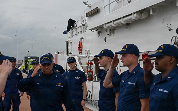A group of U.S. Coast Guard personnel are gathered on the deck of a white Coast Guard cutter, performing a formal salute ceremony. A senior officer, identifiable by rank insignia on their cap, is walking along the deck while crew members on both sides render salutes. The scene appears to be a formal inspection, boarding, or departure ceremony conducted in overcast, wet weather conditions.