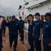 A group of U.S. Coast Guard personnel are gathered on the deck of a white Coast Guard cutter, performing a formal salute ceremony. A senior officer, identifiable by rank insignia on their cap, is walking along the deck while crew members on both sides render salutes. The scene appears to be a formal inspection, boarding, or departure ceremony conducted in overcast, wet weather conditions.