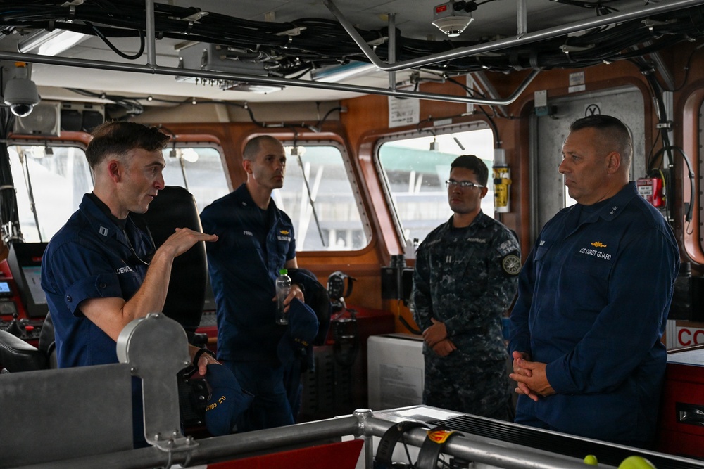 Four military personnel are gathered on the bridge/wheelhouse of a U.S. Coast Guard vessel. One officer on the left appears to be briefing or explaining something to the group, including what appears to be a senior Coast Guard officer on the right and a Navy personnel member in digital camouflage uniform. The setting is an operational vessel bridge with navigation equipment and controls visible.