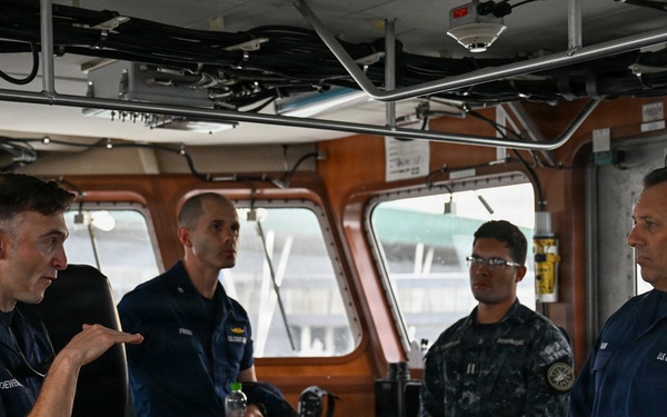Four military personnel are gathered on the bridge/wheelhouse of a U.S. Coast Guard vessel. One officer on the left appears to be briefing or explaining something to the group, including what appears to be a senior Coast Guard officer on the right and a Navy personnel member in digital camouflage uniform. The setting is an operational vessel bridge with navigation equipment and controls visible.
