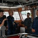 Four military personnel are gathered on the bridge/wheelhouse of a U.S. Coast Guard vessel. One officer on the left appears to be briefing or explaining something to the group, including what appears to be a senior Coast Guard officer on the right and a Navy personnel member in digital camouflage uniform. The setting is an operational vessel bridge with navigation equipment and controls visible.