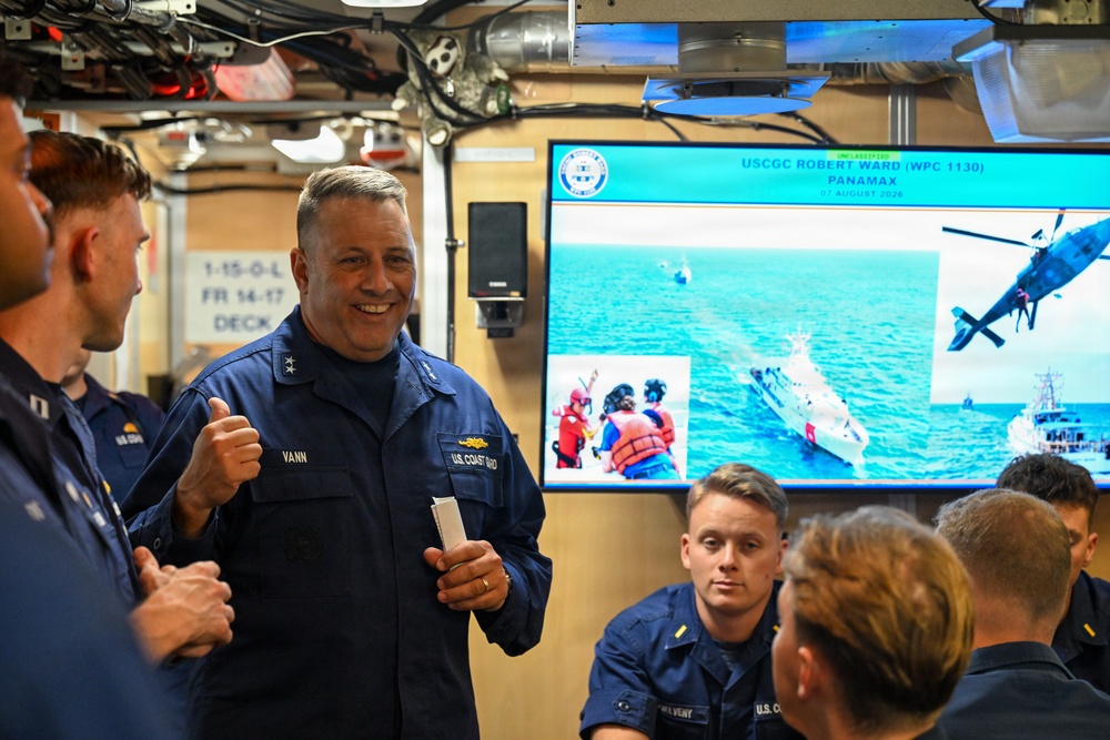 A U.S. Coast Guard officer with the name tag 'VANN' and two stars on his collar (indicating a Rear Admiral rank) is giving a thumbs-up while speaking with a group of Coast Guard personnel in what appears to be an onboard briefing room. Behind him, a large screen displays a presentation titled 'USCGC ROBERT WARD (WPC 1130) PANAMAX' dated 07 August 2026, showing maritime rescue operation imagery. The setting appears to be aboard a Coast Guard vessel during an operational briefing or visit.