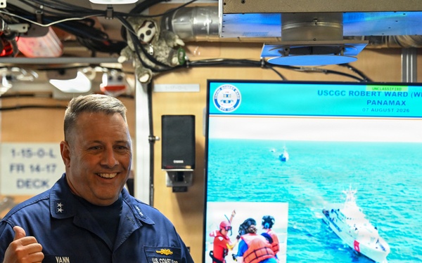 A U.S. Coast Guard officer with the name tag 'VANN' and two stars on his collar (indicating a Rear Admiral rank) is giving a thumbs-up while speaking with a group of Coast Guard personnel in what appears to be an onboard briefing room. Behind him, a large screen displays a presentation titled 'USCGC ROBERT WARD (WPC 1130) PANAMAX' dated 07 August 2026, showing maritime rescue operation imagery. The setting appears to be aboard a Coast Guard vessel during an operational briefing or visit.