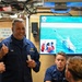A U.S. Coast Guard officer with the name tag 'VANN' and two stars on his collar (indicating a Rear Admiral rank) is giving a thumbs-up while speaking with a group of Coast Guard personnel in what appears to be an onboard briefing room. Behind him, a large screen displays a presentation titled 'USCGC ROBERT WARD (WPC 1130) PANAMAX' dated 07 August 2026, showing maritime rescue operation imagery. The setting appears to be aboard a Coast Guard vessel during an operational briefing or visit.