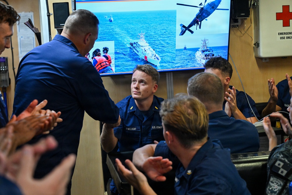 A U.S. Coast Guard ceremony or recognition event is taking place aboard what appears to be a Coast Guard vessel or facility. A senior officer is shaking hands with a seated Coast Guard member identified by name tape as 'O'MELVENY,' while surrounding personnel applaud. A large screen in the background displays Coast Guard operational imagery including vessels and a helicopter.