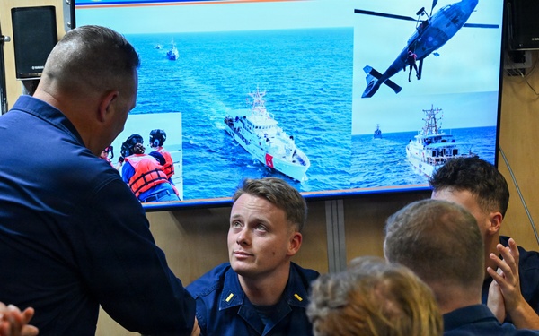 A U.S. Coast Guard ceremony or recognition event is taking place aboard what appears to be a Coast Guard vessel or facility. A senior officer is shaking hands with a seated Coast Guard member identified by name tape as 'O'MELVENY,' while surrounding personnel applaud. A large screen in the background displays Coast Guard operational imagery including vessels and a helicopter.