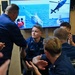 A U.S. Coast Guard ceremony or recognition event is taking place aboard what appears to be a Coast Guard vessel or facility. A senior officer is shaking hands with a seated Coast Guard member identified by name tape as 'O'MELVENY,' while surrounding personnel applaud. A large screen in the background displays Coast Guard operational imagery including vessels and a helicopter.