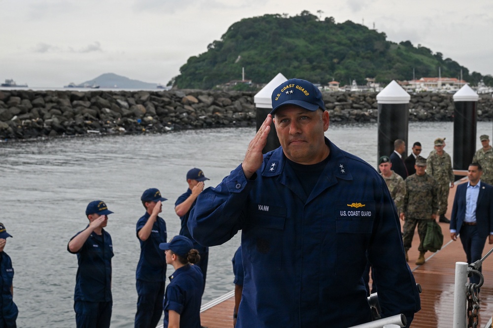 A U.S. Coast Guard officer with the name tag 'VANN' and two-star rank insignia (Rear Admiral) is rendering a salute on a wet dock or pier. Behind him, a line of Coast Guard personnel in blue uniforms are also saluting, while military personnel in camouflage uniforms and civilians are visible in the background. The setting appears to be a coastal harbor with a rocky breakwater and a green, forested island visible in the background.