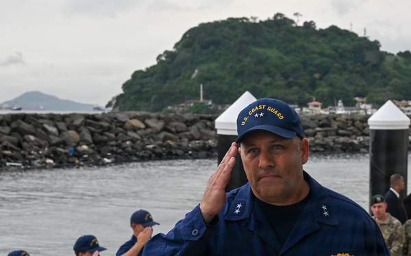 A U.S. Coast Guard officer with the name tag 'VANN' and two-star rank insignia (Rear Admiral) is rendering a salute on a wet dock or pier. Behind him, a line of Coast Guard personnel in blue uniforms are also saluting, while military personnel in camouflage uniforms and civilians are visible in the background. The setting appears to be a coastal harbor with a rocky breakwater and a green, forested island visible in the background.