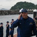 A U.S. Coast Guard officer with the name tag 'VANN' and two-star rank insignia (Rear Admiral) is rendering a salute on a wet dock or pier. Behind him, a line of Coast Guard personnel in blue uniforms are also saluting, while military personnel in camouflage uniforms and civilians are visible in the background. The setting appears to be a coastal harbor with a rocky breakwater and a green, forested island visible in the background.