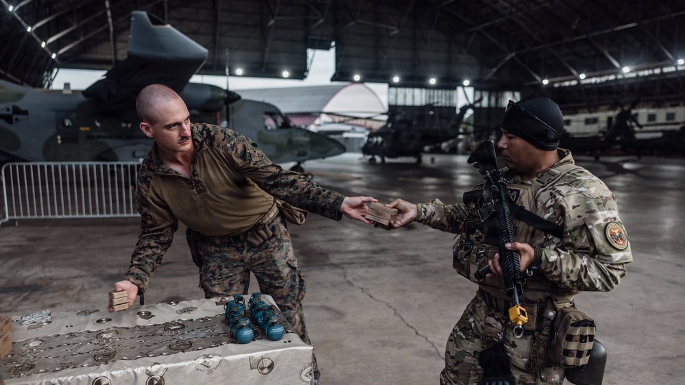 Two military personnel are inside a large aircraft hangar exchanging what appears to be a small box or package across a table. The table holds several blue spherical objects, likely training grenades or similar equipment. Multiple military aircraft are visible in the background of the hangar.