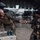 PANAMAX-26: V1/6, Partner forces conduct simulated air raid Two military personnel are inside a large aircraft hangar exchanging what appears to be a small box or package across a table. The table holds several blue spherical objects, likely training grenades or similar equipment. Multiple military aircraft are visible in the background of the hangar.