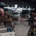 Two military personnel are inside a large aircraft hangar exchanging what appears to be a small box or package across a table. The table holds several blue spherical objects, likely training grenades or similar equipment. Multiple military aircraft are visible in the background of the hangar.