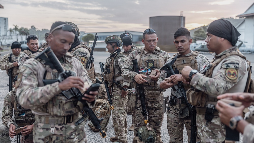 A group of military or law enforcement personnel in multicam/camouflage uniforms are gathered on what appears to be a tarmac or airfield apron at dusk. The personnel are equipped with tactical gear, body armor, and rifles, and appear to be conducting pre-mission equipment checks or briefings. Patches and insignia visible on uniforms suggest a multinational or joint operation context.