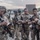 PANAMAX-26: V1/6, Partner forces conduct simulated air raid A group of military or law enforcement personnel in multicam/camouflage uniforms are gathered on what appears to be a tarmac or airfield apron at dusk. The personnel are equipped with tactical gear, body armor, and rifles, and appear to be conducting pre-mission equipment checks or briefings. Patches and insignia visible on uniforms suggest a multinational or joint operation context.