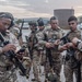 A group of military or law enforcement personnel in multicam/camouflage uniforms are gathered on what appears to be a tarmac or airfield apron at dusk. The personnel are equipped with tactical gear, body armor, and rifles, and appear to be conducting pre-mission equipment checks or briefings. Patches and insignia visible on uniforms suggest a multinational or joint operation context.