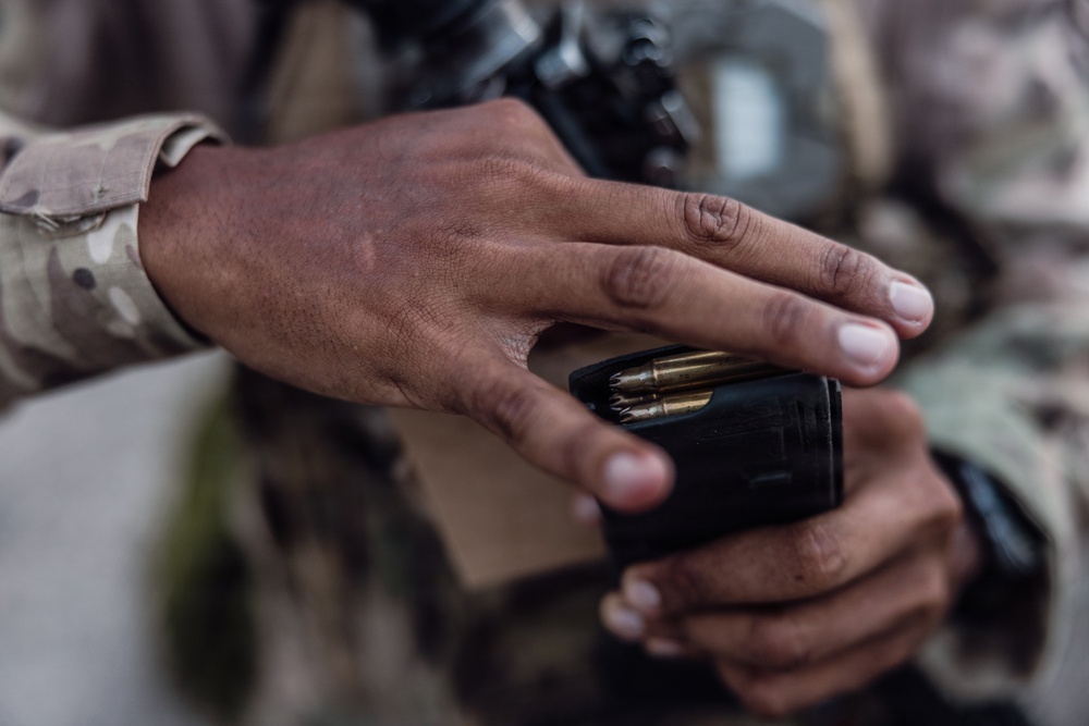 A close-up photograph showing the hands of a person wearing camouflage military uniform handling a black firearm magazine loaded with brass-cased rifle cartridges. The image focuses on the act of loading or inspecting ammunition. A firearm is partially visible in the blurred background.