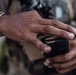 A close-up photograph showing the hands of a person wearing camouflage military uniform handling a black firearm magazine loaded with brass-cased rifle cartridges. The image focuses on the act of loading or inspecting ammunition. A firearm is partially visible in the blurred background.