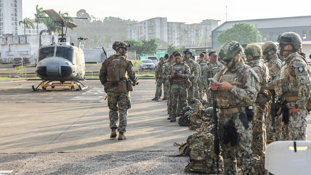 A group of military personnel in camouflage uniforms are assembled on a tarmac or helipad area near a UH-1 'Huey' utility helicopter. Multiple soldiers wearing tactical gear, helmets, and carrying weapons are visible, with some bearing 'MARINA' (Navy) markings on their vests, suggesting Mexican Naval Infantry involvement. The scene appears to be a pre-mission briefing or staging operation in an urban tropical environment.